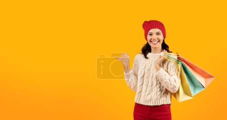 Photo for Winter Shopping And Sales. Happy Customer Lady In Red Knitwear Holding Shopper Bags And Pointing Thumb Finger Aside, Showing Free Space On Yellow Studio Background. Panorama - Royalty Free Image