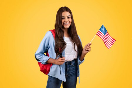 Foto de Mujer adolescente caucásica positiva, con bandera de Estados Unidos, combina patriotismo con academia, aislada sobre fondo amarillo. Estilo de vida, orgullo nacional y estudio de intercambio, aprender inglés - Imagen libre de derechos