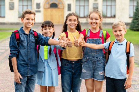 Photo for Group of happy kids wearing backpacks showing thumbs up while posing outdoors, smiling pupils standing against school building, recommending educational programs, enjoying study together - Royalty Free Image