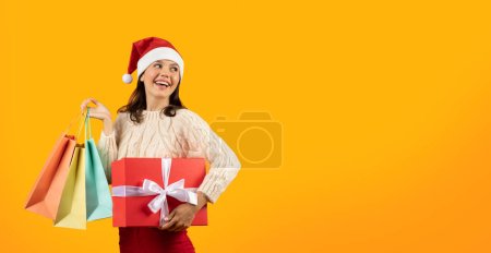 Foto de Concepto de compras de Navidad. Mujer alegre en el sombrero de Santa Claus sosteniendo bolsas de comprador y caja de regalo en el fondo del estudio amarillo. Feliz cliente comprando regalos para Navidad, espacio vacío, panorama - Imagen libre de derechos