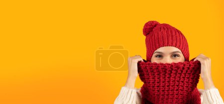 Téléchargez les photos : Jeune femme portant un chapeau tricoté rouge et une écharpe se cachant du froid hivernal sur fond jaune studio, couvrant le visage avec une écharpe chaude regardant la caméra, Panorama, Espace libre - en image libre de droit