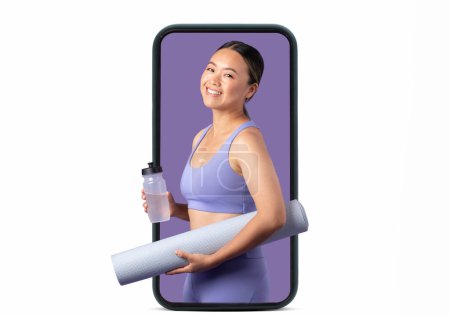 Foto de Deportiva mujer asiática en ropa deportiva, sosteniendo esterilla de yoga y botella de agua, aparece lista para el entrenamiento, mostrada digitalmente en la pantalla del teléfono inteligente contra el fondo del estudio blanco - Imagen libre de derechos