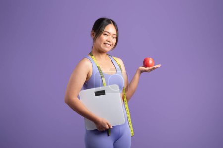 Foto de Mujer asiática sonriente en ropa deportiva sosteniendo escamas en una mano y manzana roja en la otra, promoviendo una dieta equilibrada sobre fondo púrpura estudio, espacio libre - Imagen libre de derechos