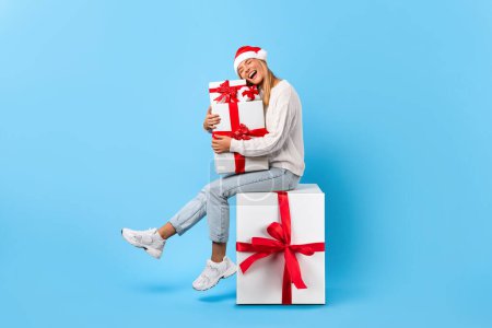 Téléchargez les photos : Femme excitée posant avec de nombreux cadeaux de Noël et assis sur une grande boîte cadeau, dame portant le chapeau de Père Noël, posant sur fond bleu studio. Ventes saisonnières et célébration du Nouvel An - en image libre de droit