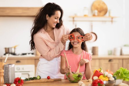 Foto de Alegre mamá y niña haciendo ensalada de verduras y divertirse, mami sosteniendo rodajas de pimienta como gafas cerca de ojos hijas, engañando en la cocina moderna en el interior - Imagen libre de derechos