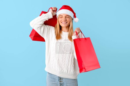Téléchargez les photos : Concept de ventes saisonnières. Femme excitée dans le chapeau du Père Noël tenant des sacs à provisions, dame étonnée heureuse de nouveaux achats, fond bleu studio - en image libre de droit