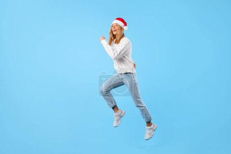 Photo for Christmas rush. Happy lady in Santa hat jumping on blue background, in motion shot of excited woman running to buy presents, full length shot, copy space - Royalty Free Image