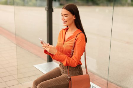 Photo for Casual young lady with smartphone sitting at the bus or tram stop and using city travel application outdoor, paying for ticket ride online via cellphone in urban area. Transportation - Royalty Free Image
