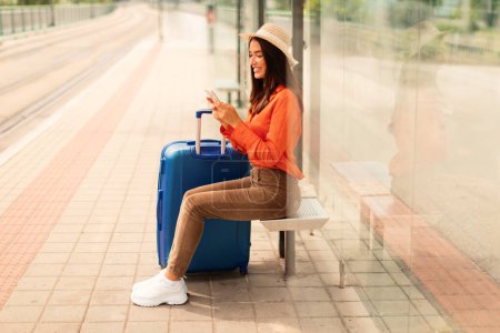 Photo for Passenger lady using cellphone monitoring traffic schedule via application sitting at tram stop outside, waiting with suitcase luggage and paying for ride online. Urban transportation - Royalty Free Image