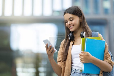 Photo for Smiling pretty young woman student using app on smartphone, navigates through an educational app on her smartphone, blending learning and technology at a bustling university campus, copy space - Royalty Free Image