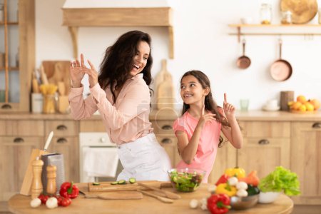 Foto de Madre e hija positiva bailando y disfrutando mientras preparan la cena en la cocina moderna interior, divirtiéndose y compartiendo momentos alegres durante la preparación de ensaladas. Ocio familiar y nutrición - Imagen libre de derechos
