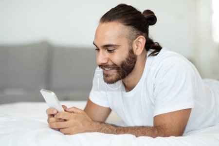 Téléchargez les photos : Joyeux bel homme caucasien en pyjama couché sur le lit à la maison, en utilisant un smartphone et souriant, vérifier les médias sociaux tout en se refroidissant seul le week-end, copier l'espace. Loisirs et gadgets - en image libre de droit