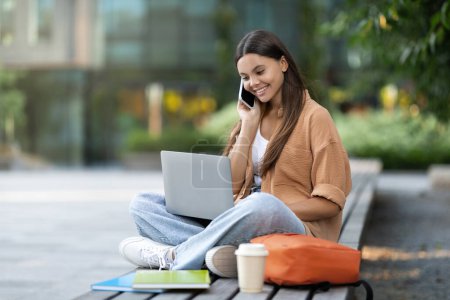 Photo for A happy young woman student at an empty university campus converses on the phone and types on her laptop, likely discussing online coursework or coordinating a study group, copy space - Royalty Free Image