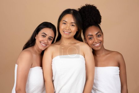 Téléchargez les photos : Trois femmes multiraciales, chacune ornée de serviette, posent magnifiquement sur fond de studio beige, créant un tableau serein de beauté, de simplicité et d'unité multiculturelle - en image libre de droit