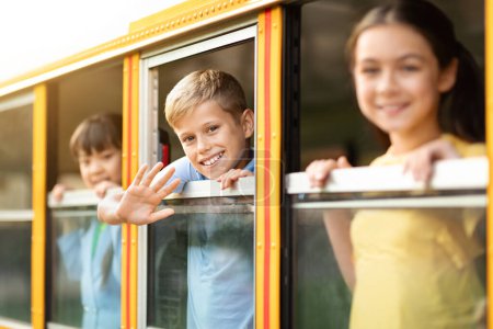Foto de Niños alegres mirando desde el autobús escolar, mirando por la ventana y sonriendo a la cámara, niños multiétnicos felices listos para el viaje juntos, disfrutando del viaje, enfoque selectivo en el niño preadolescente saludando la mano - Imagen libre de derechos