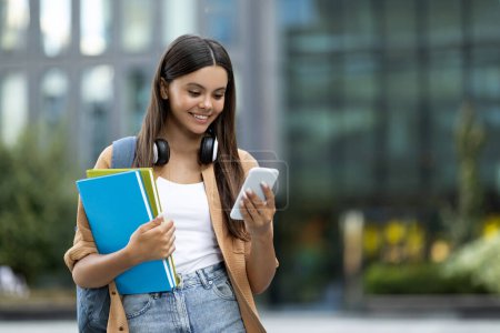 Photo for Young woman student texting on smartphone at campus, copy space. Positive cheerful lady with backpack and notebooks in her hand walking by street, using phone, copy space. Communication - Royalty Free Image