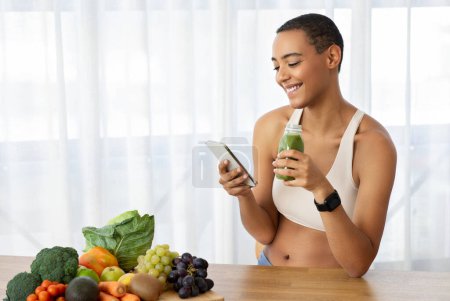 Photo for Cheerful young latin lady in sportswear, drink jar of fresh smoothie, juice, watch video on phone on kitchen interior with fresh vegetables. Diet at home, healthy food, fit app - Royalty Free Image