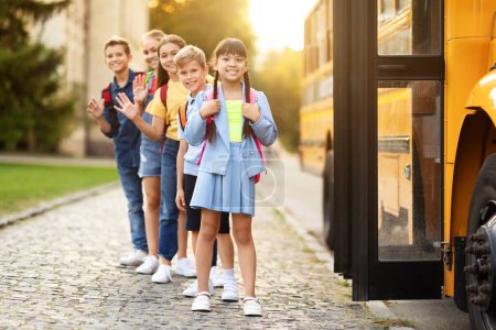 Foto de Niños con mochilas de pie junto al autobús escolar, niños multiétnicos felices anticipando un cómodo viaje a casa después de un día escolar activo, mirando y saludando con las manos a la cámara, longitud completa - Imagen libre de derechos