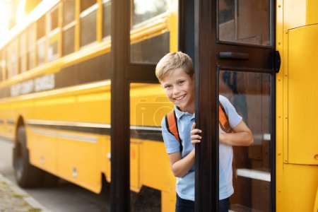 Foto de Feliz niño sonriente de pie en la puerta del autobús escolar, niño alegre con mochila entrar en el vehículo, listo para un divertido día de aprender y jugar con amigos, mirando a la cámara, espacio de copia - Imagen libre de derechos