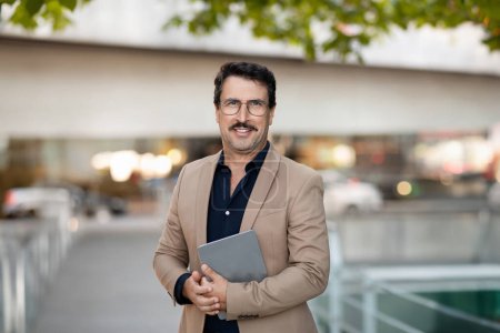 Photo for Handsome middle aged man confidently holds a digital tablet, navigating its content while standing on a bustling city street, representing a fusion of modern tech and everyday life - Royalty Free Image