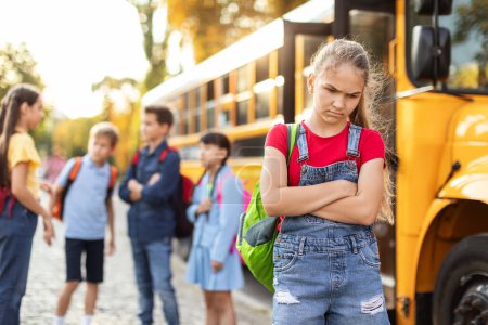 Photo for Upset preteen girl standing alone near yellow school bus while classmates happy chatting on background, offended female child folding arms and frowning, feeling loneliness and social exclusion - Royalty Free Image