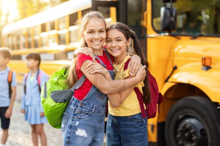 Téléchargez les photos : Enfants joyeux debout à côté du bus scolaire jaune, profitant de moments heureux ensemble avant de rentrer à la maison, deux pré-adolescentes positives avec des sacs à dos embrassant et souriant à la caméra, espace libre - en image libre de droit