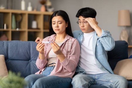Photo for Unhappy Japanese couple holding a negative pregnancy test result, sitting on sofa, hugging and providing support amid this difficult moment at home. Reproductive and childbirth healthcare struggle - Royalty Free Image