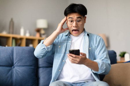 Foto de Emocional chico japonés que trata con el problema del teléfono celular en la sala de estar, mirando gadget teléfono en shock mientras lee noticias o mensajes desagradables, sentado en el sofá en casa y tocando la cabeza - Imagen libre de derechos