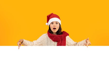 Téléchargez les photos : Offre de Noël. Jeune dame étonnante montrant l'affiche en papier avec l'espace vide pour le texte, pointant du doigt annonçant des ventes de Noël, portant le chapeau de Santa sur fond jaune de studio, maquette - en image libre de droit