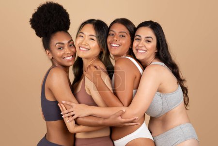 Multirassische Frauen strahlen natürliche Schönheit aus, posieren selbstbewusst in Unterwäsche und schaffen eine harmonische, körperbetonte Szene vor beigem Studiohintergrund