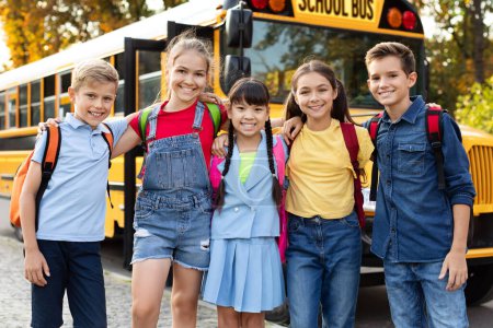 Foto de Niños sonrientes felices esperando junto al autobús escolar amarillo, grupo de niños alegres con mochilas listas para ir a casa después del día de aprendizaje y diversión, alumnos positivos abrazando y mirando a la cámara - Imagen libre de derechos