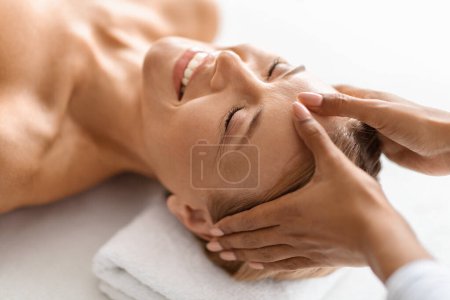 Téléchargez les photos : Femme d'âge moyen détendue bénéficiant d'un massage apaisant de la tête à l'acupression au salon de spa de luxe, femme calme allongée sur la table et souriante, ayant un traitement de bien-être facilitant la libération de tension et la sérénité - en image libre de droit