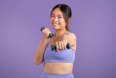 Téléchargez les photos : Femme asiatique énergique en vêtements de sport s'engage avec enthousiasme dans l'exercice à l'aide d'haltères à main, démontrant fitness et bien-être sur fond violet studio - en image libre de droit
