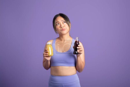 Téléchargez les photos : Femme asiatique sportive hésite tout en faisant un choix entre smoothie sain et cola malsain, présentant dilemme nutritionnel, sur fond de studio violet - en image libre de droit