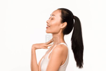 Photo for Elegant profile of Asian woman with sleek long black hair tied in a ponytail, posing with eyes closed, showcasing serene expression, touches her neck with grace, white background - Royalty Free Image