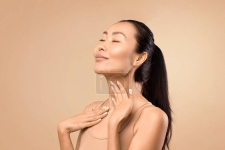 Téléchargez les photos : Femme asiatique d'âge moyen avec les yeux fermés touchant doucement son cou, exprimant sa satisfaction avec son état de peau, studio tourné avec un fond beige - en image libre de droit
