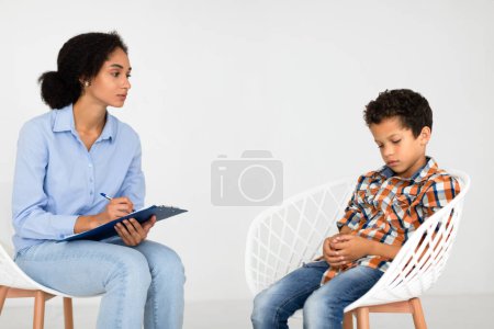 Photo for Psychotherapist woman noting as distressed boy sits lost in thoughts indoors over white wall background. Young childs mental challenges and the support of professional counseling concept - Royalty Free Image