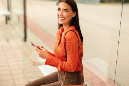 Photo for Cheerful passenger young woman holds mobile phone, booking tram ride tickets while waiting sitting at city stop outside. European lady texting awaiting public transport in urban area - Royalty Free Image