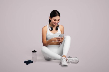Photo for Fit young woman wearing headphones sits relaxed, engaging with her smartphone after workout session, set against light background, full length, free space - Royalty Free Image