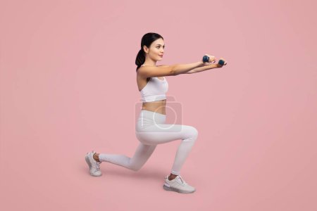 Photo for Focused young woman in white workout outfit confidently performs a lunge while extending dumbbells forward, set against soft pink backdrop, full length, free space - Royalty Free Image