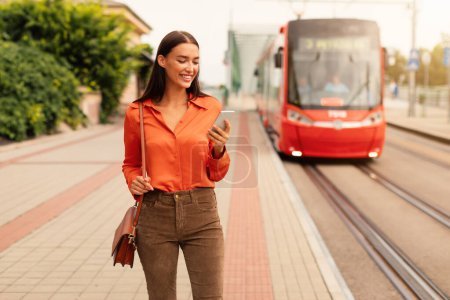 Photo for Lady passenger with smartphone purchases tram ride tickets at outdoor city stop, while red streetcar arriving at station in urban area. Mobile application for easy urban commuting - Royalty Free Image