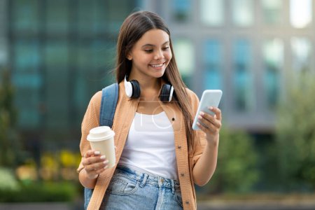 Téléchargez les photos : Engagé joyeux jeune étudiante shopping en ligne, en utilisant son smartphone, tout en marchant par ensoleillé, campus universitaire dynamique, incarnant la commodité numérique, boire du café à emporter - en image libre de droit