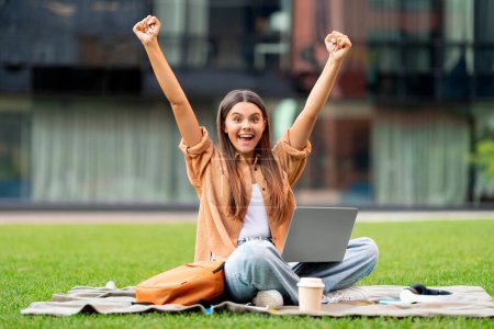 Photo for Emotional woman student with laptop on her lap raising hands up, sitting on green grass at university campus, have break between lessons, checking opportunities online, passed exam - Royalty Free Image