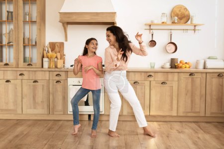 Téléchargez les photos : Joyeux jeune maman et sa fille dansant dans la cuisine de la maison, s'amusant et partageant moment de famille ludique. Plan complet. Mère et enfant fille faisant des mouvements drôles à la musique - en image libre de droit