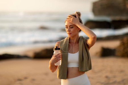 Téléchargez les photos : Femme européenne tenant une bouteille d'eau et touchant le front, fatiguée après l'entraînement, debout sur une plage de sable fin au coucher du soleil, entourée d'un paysage marin serein - en image libre de droit