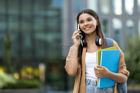 Photo for Happy cheerful pretty young woman student walking by university campus, talking on phone with friend, making plans, holding notebooks in her hand, carrying backpack, looking at copy space - Royalty Free Image
