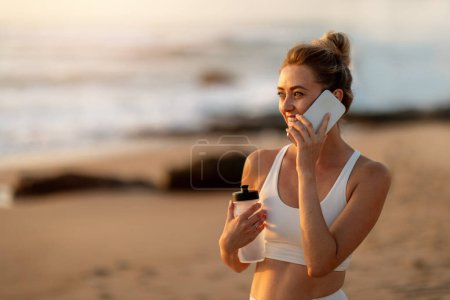 Téléchargez les photos : Femme européenne se tient au bord de la mer, parlant sur son téléphone portable après une séance de yoga au coucher du soleil sur la plage, faisant une pause après l'entraînement, espace libre - en image libre de droit