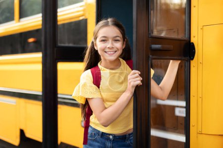 Foto de Chica feliz sonriente de pie en la puerta del autobús escolar, niña alegre con mochila que entra en el vehículo, listo para un día divertido de aprender y jugar con amigos, mirando a la cámara, espacio de copia - Imagen libre de derechos