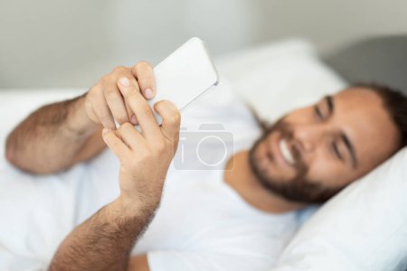 Téléchargez les photos : Un gars souriant utilisant un téléphone portable naviguant sur Internet et envoyant des SMS sur son téléphone allongé dans son lit à la maison. Heureux homme millénaire avec Smartphone réseautage dans les médias sociaux. Nouvelle application mobile Advert Concept - en image libre de droit