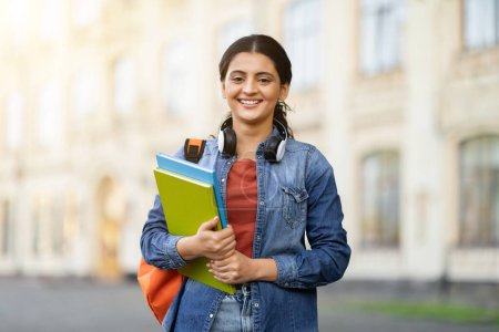 Téléchargez les photos : Joyeux jeune étudiante indienne posant à l'université ou au campus de l'université à l'extérieur, portant un sac à dos et des livres de cours, souriant à la caméra. Concept d'éducation, espace de copie - en image libre de droit